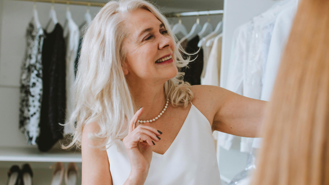 A woman with long gray hair and a white dress smiles while standing in front of an open wardrobe, selecting outfits to refresh her retirement wardrobe.